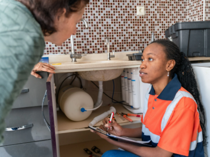 Person kneeling under a sink explains plumbing to another person in a kitchen or bathroom, holding a pen and clipboard