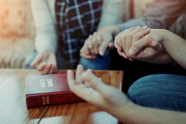 A group of people are sitting at a table holding hands in front of a bible.