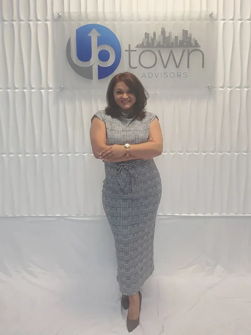 A person stands smiling with arms crossed in front of a white wall featuring the Uptown business logo.