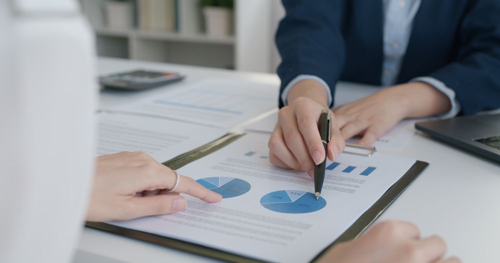 Two people in professional attire discuss data on a document containing pie charts and bar graphs at an office desk.