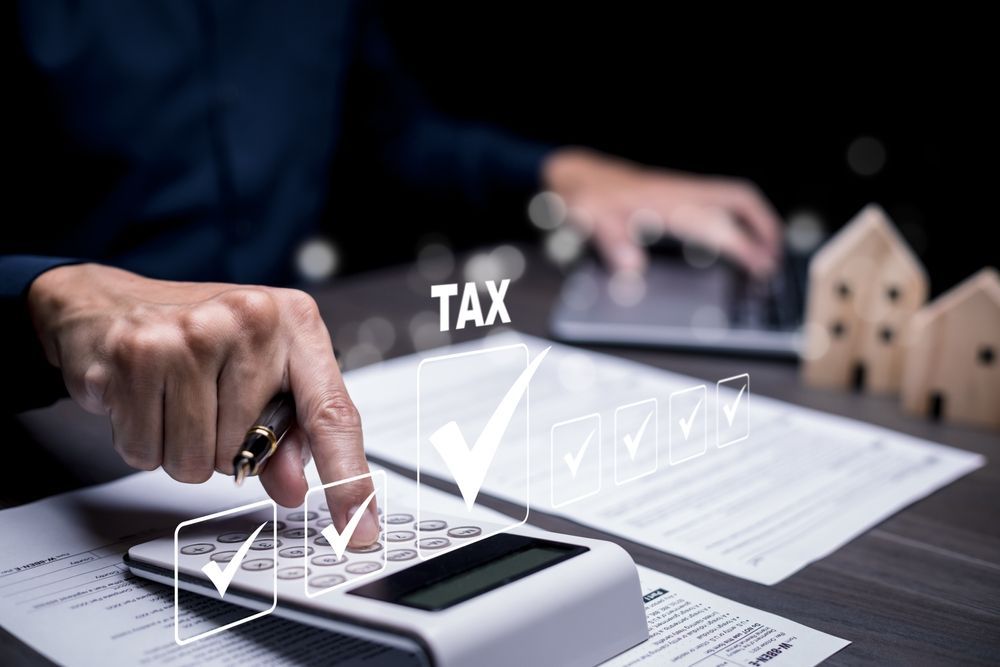 A person uses a calculator over tax documents, with a digital checkmark graphic overlay and small wooden house models nearby.