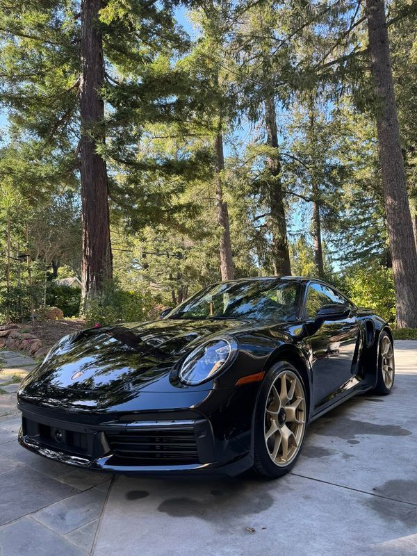 A black porsche 911 is parked in a driveway in front of trees.