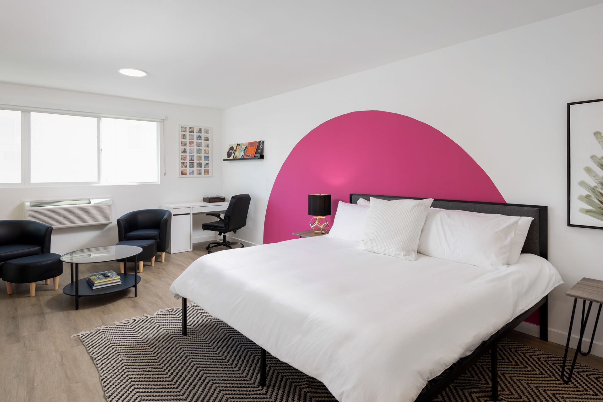 A modern bedroom featuring a bed with white bedding, a pink semicircular wall mural, a desk, and two black armchairs.