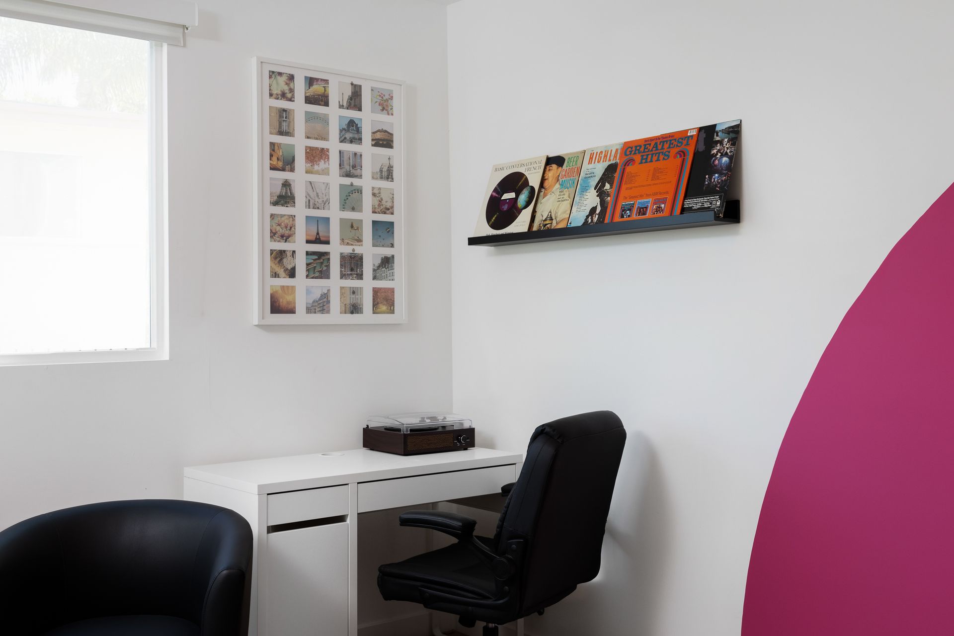 A home office corner with a white desk, a black office chair, a framed photo grid, and a shelf displaying vinyl records.