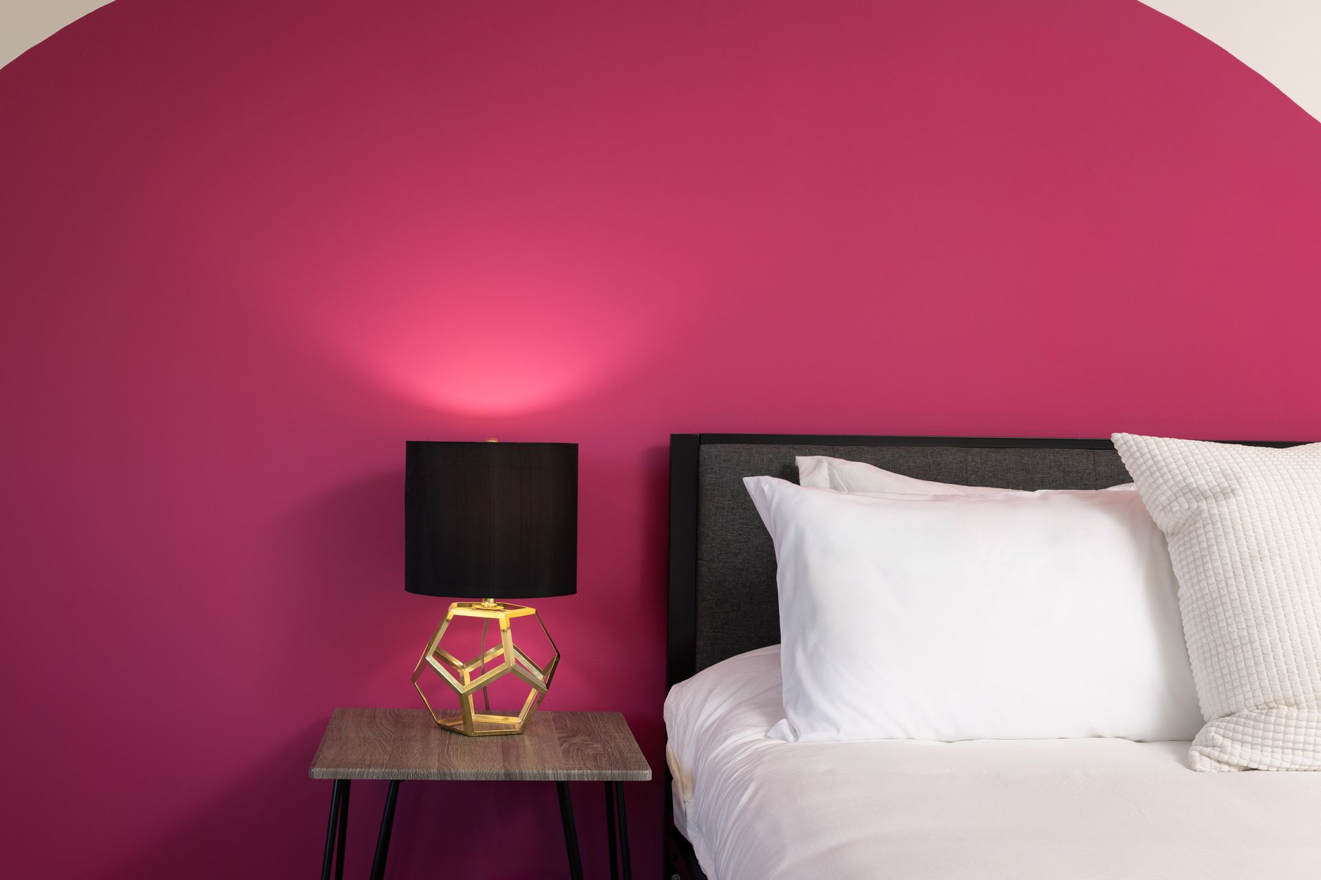 A bed with white linens and a geometric lamp on a side table against a pink wall.