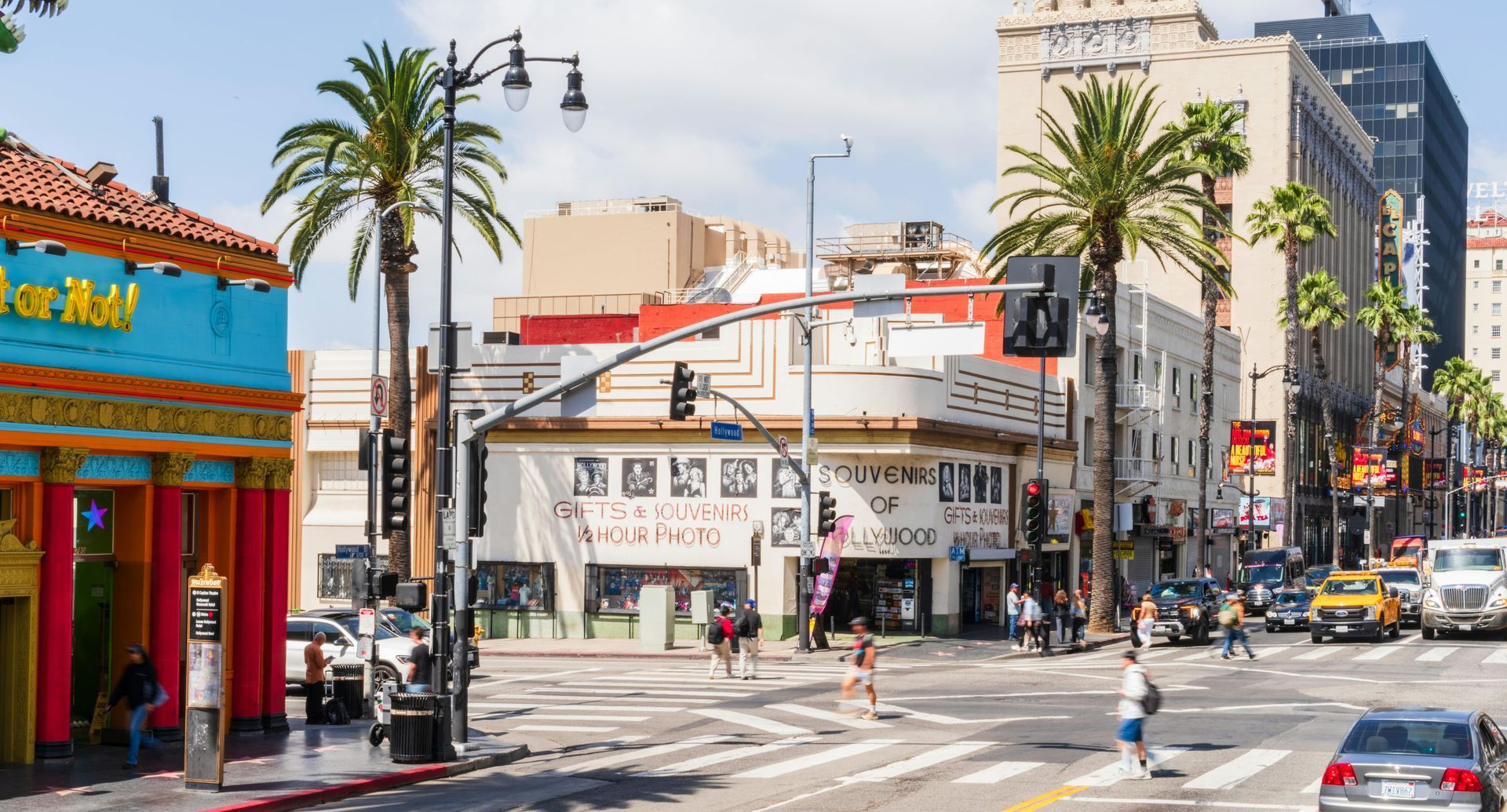 A sunny street corner in Hollywood with a vibrant blue building, palm trees, and people walking across a crosswalk.