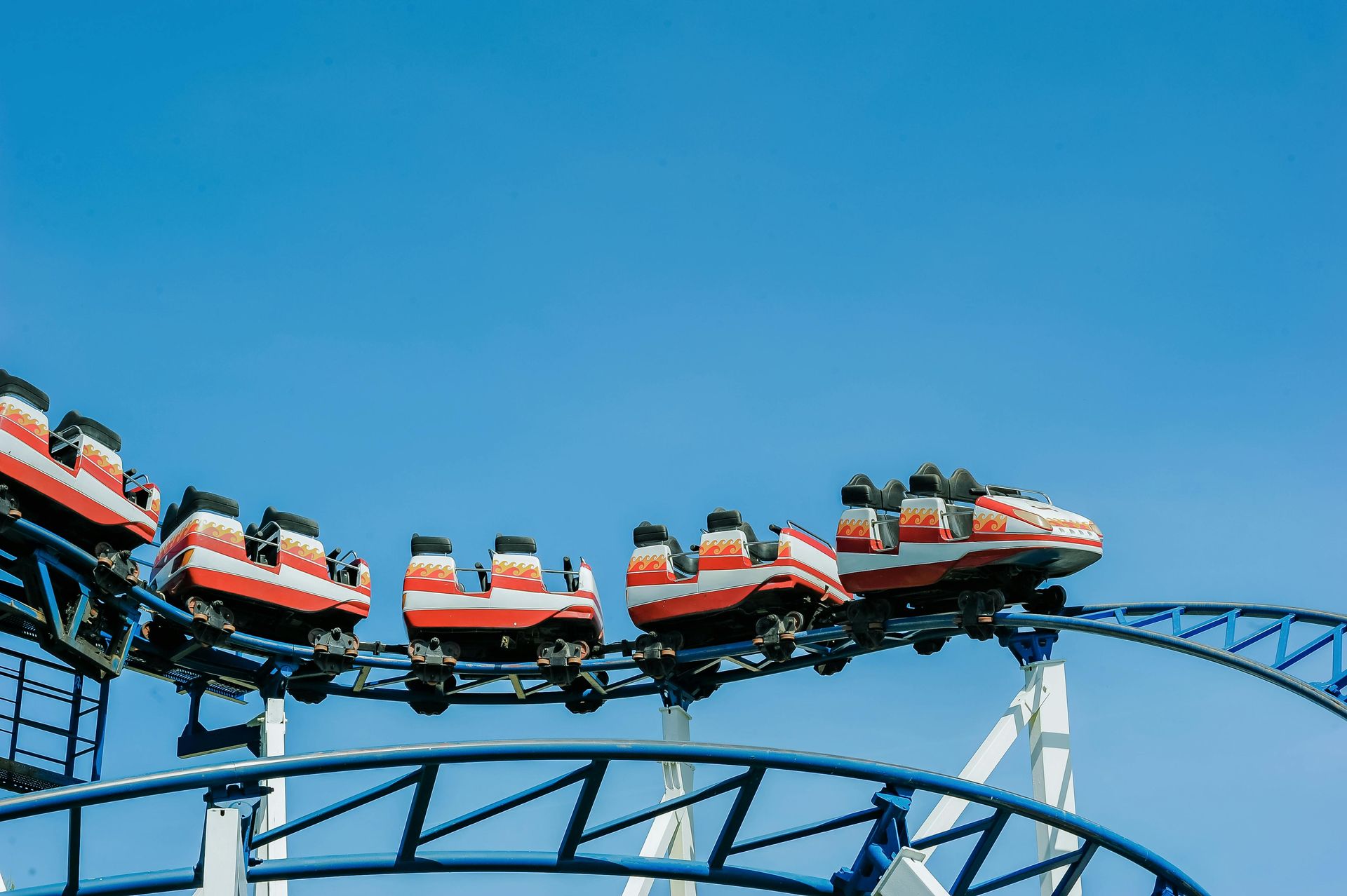 Red and white roller coaster cars travel along a blue steel track against a clear blue sky.