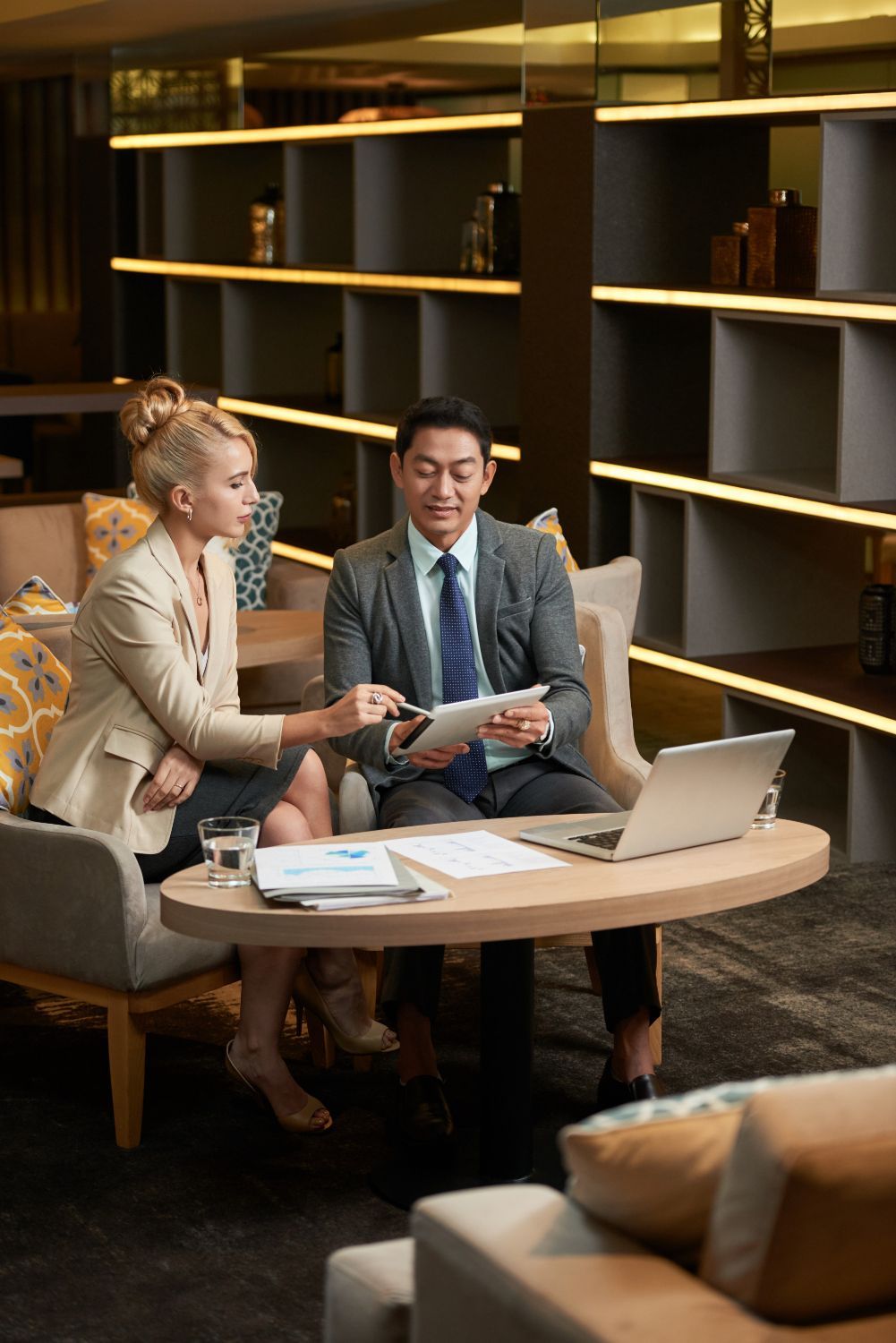 Two people reviewing documents at a table. One points to a tablet. Dark, modern setting.