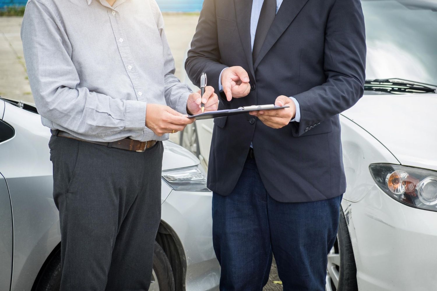 Two men by damaged cars, one signing a document held by the other, outdoors.