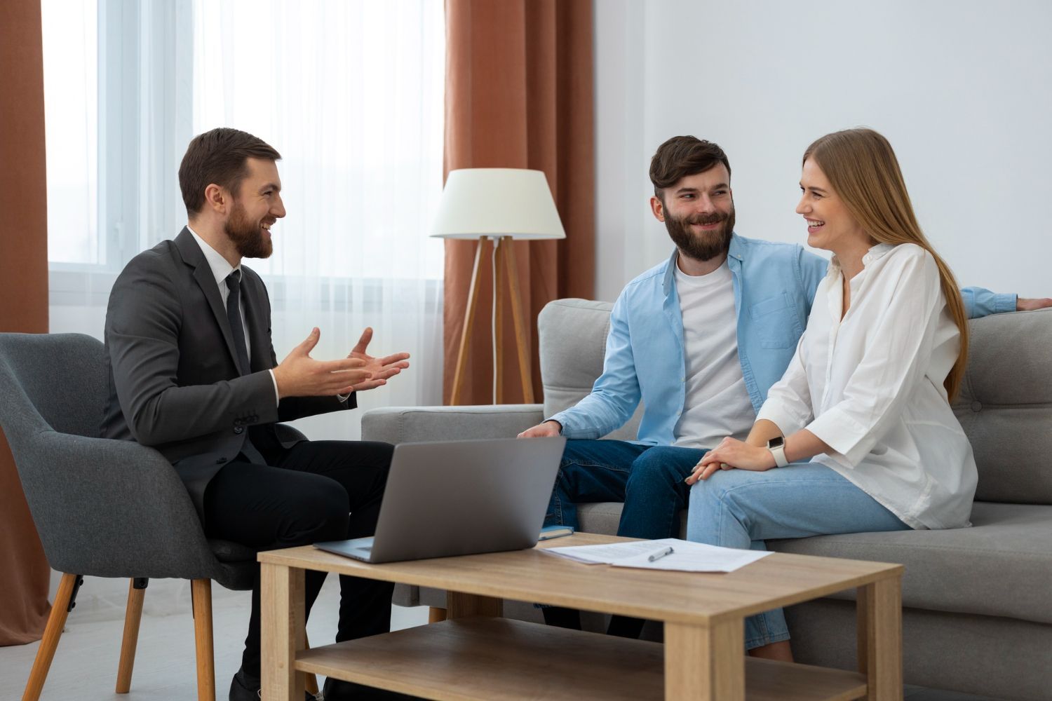 Man in suit talking to a couple on a couch; laptop and papers on coffee table.