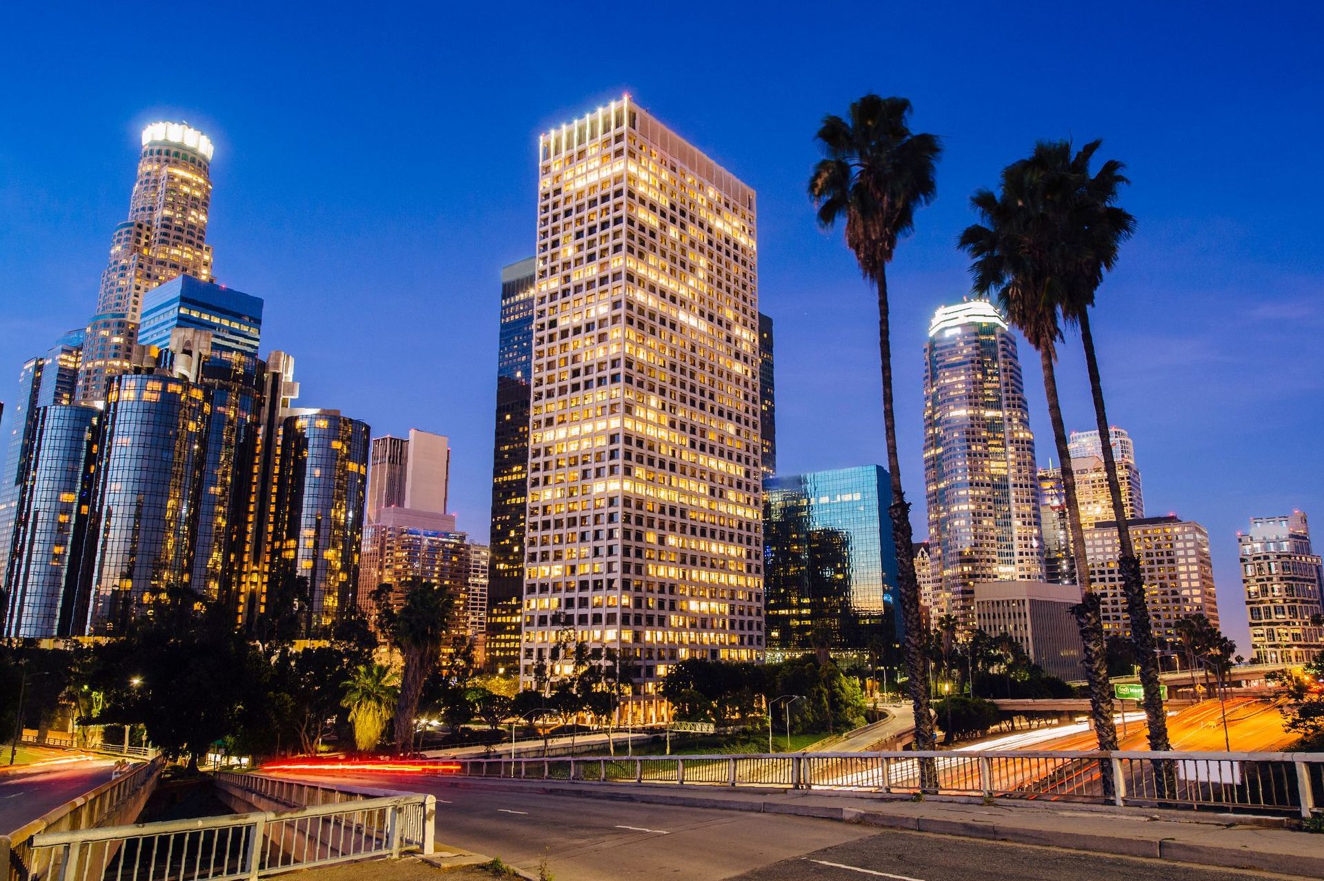 Downtown Los Angeles skyline at dusk, illuminated buildings, palm trees along a road.