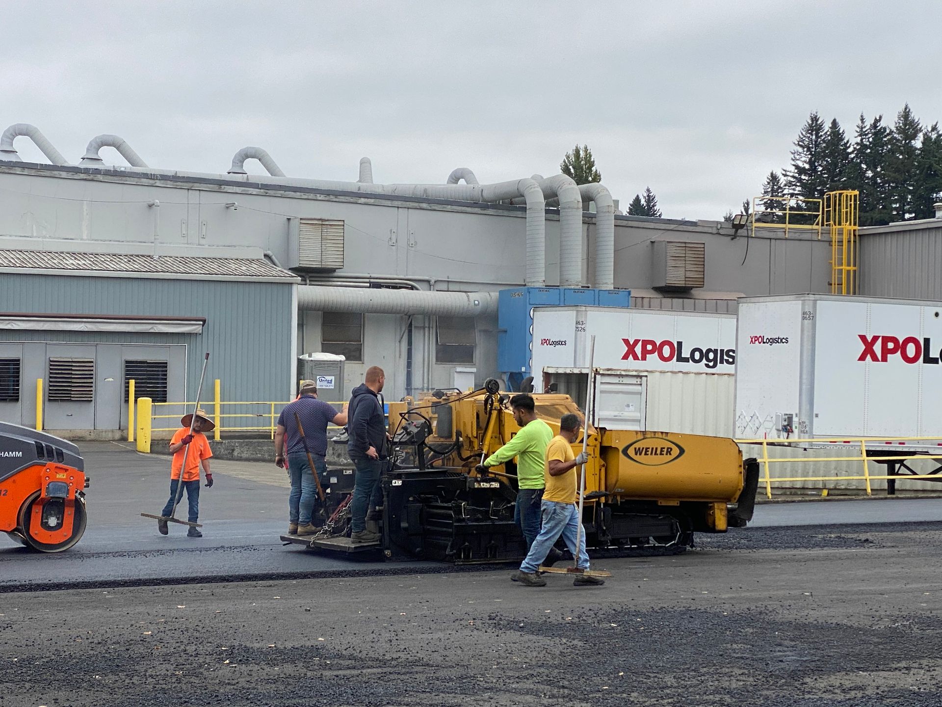 Asphalt paving crew at work, using a paver with several workers and a roller, near a warehouse.