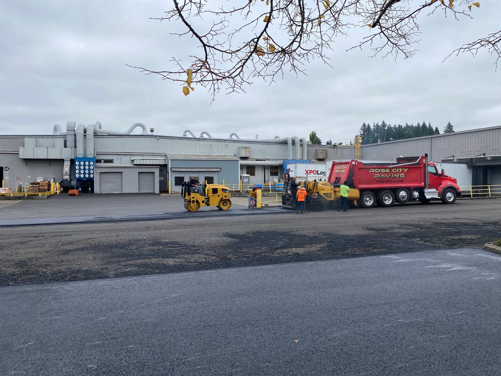 Asphalt paving in progress: Roller compacts fresh black asphalt near a red dump truck and building under cloudy sky.