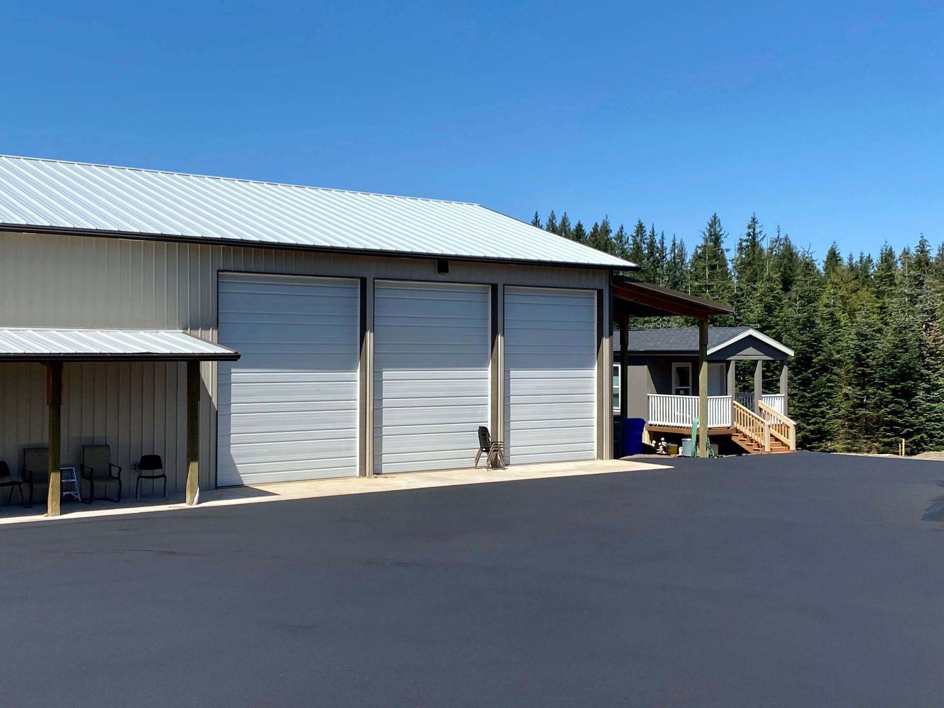 Building with three garage doors and small house in a paved lot, under a blue sky.