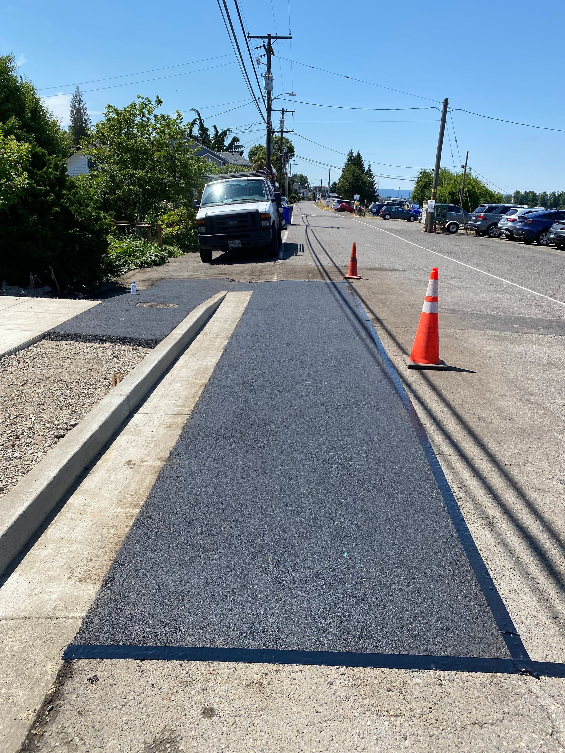 Paved road with dark gravel patches. An orange cone stands near the road. A white van is parked nearby.
