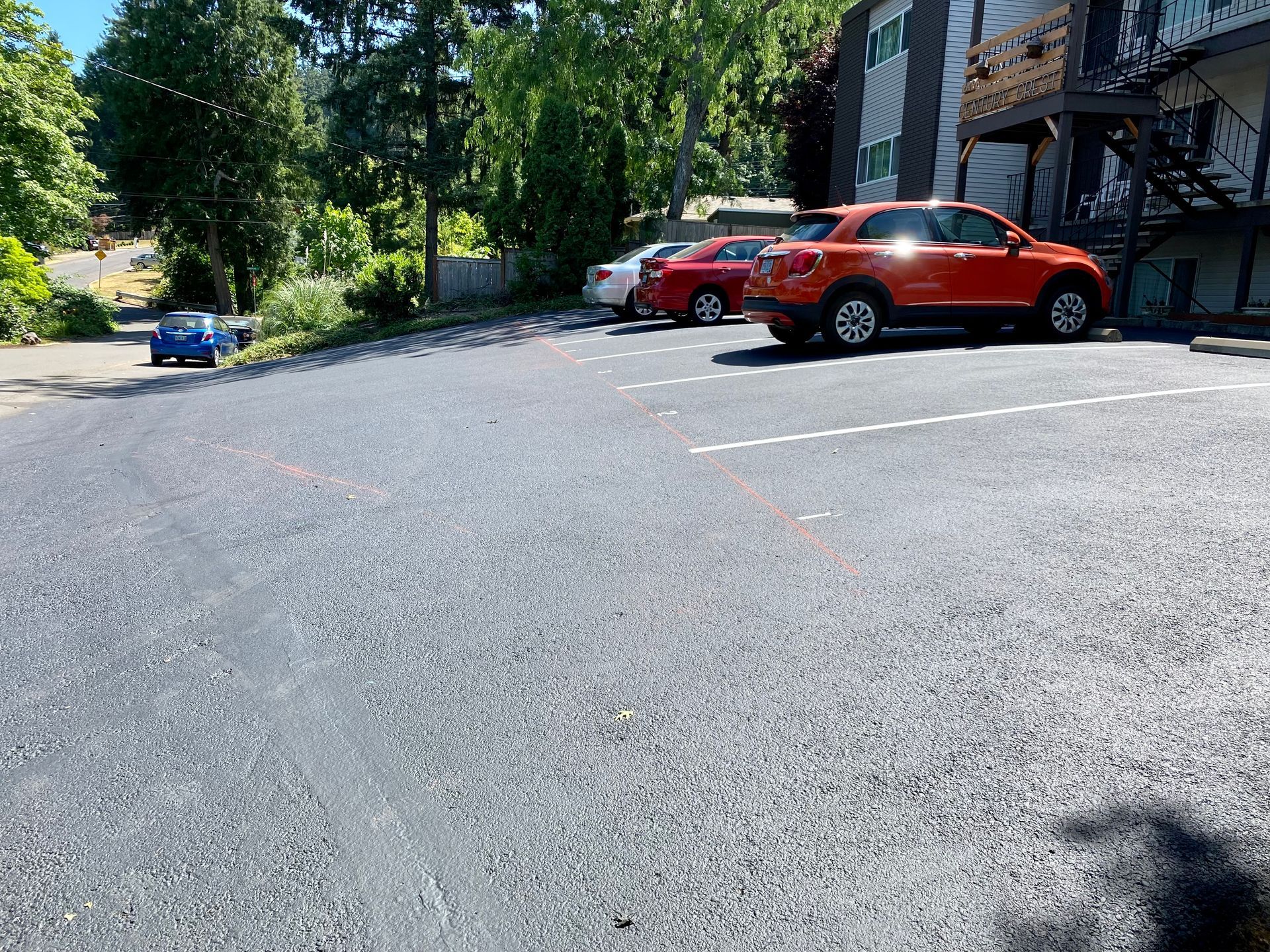 Paved parking lot with cars parked. Red and white cars, trees in the background, apartment building on the right.