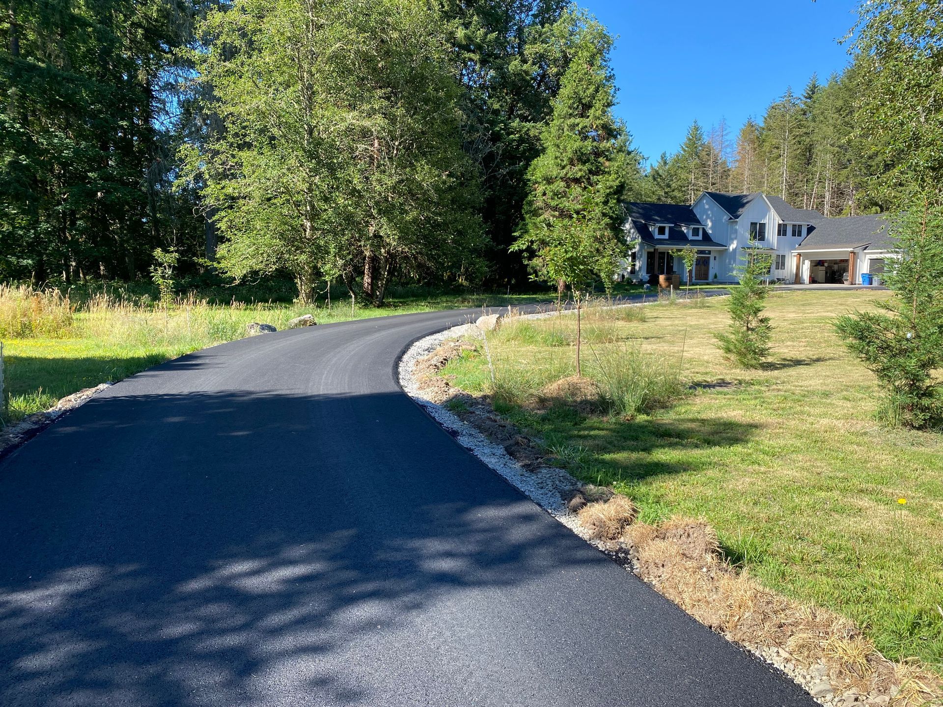 Asphalt driveway curves toward a white house surrounded by trees and grass on a sunny day.