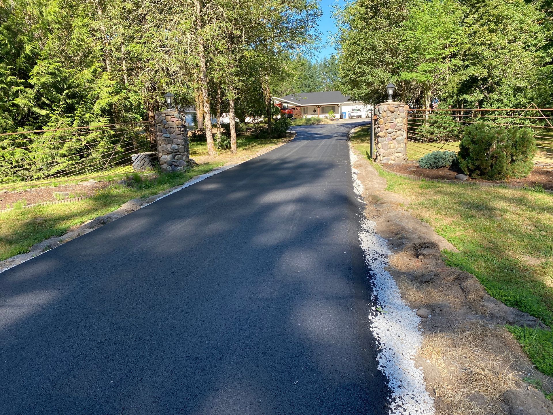 Paved driveway leading toward a house, flanked by stone columns and trees.