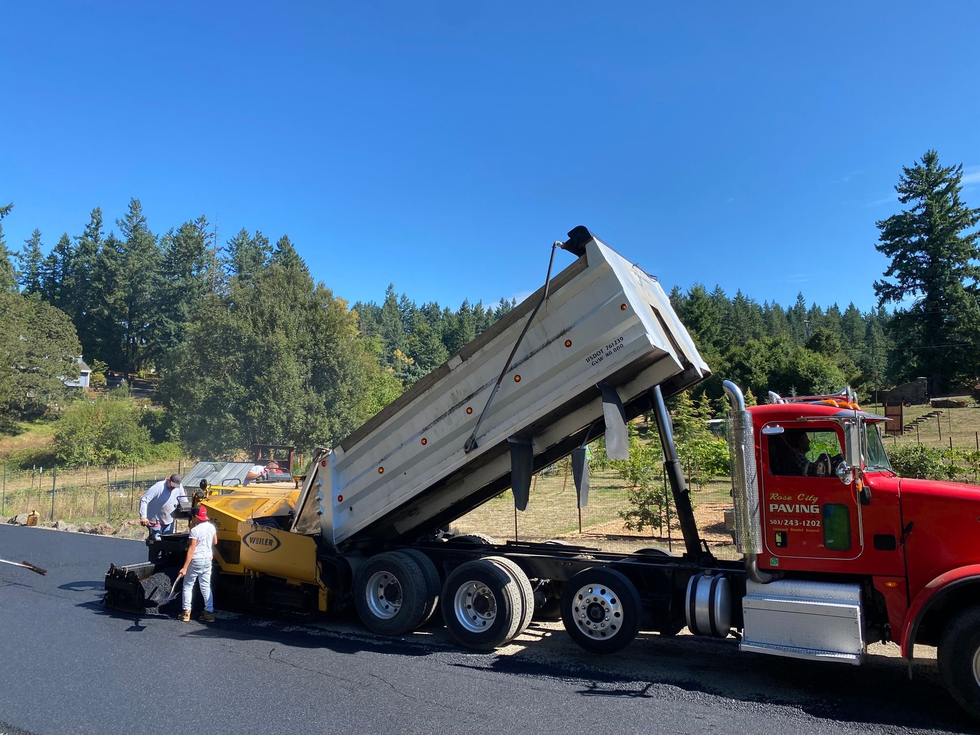 Red dump truck unloading asphalt into a paving machine on a road, two workers nearby, trees in background.
