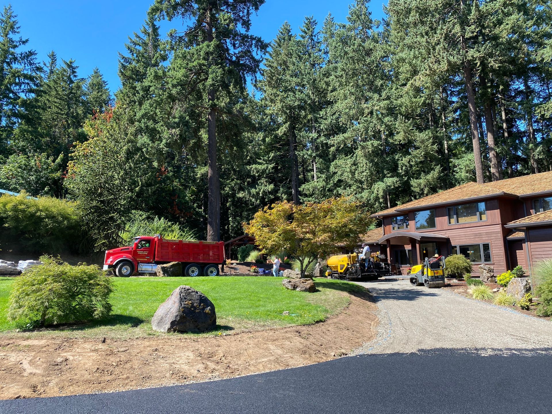 Red dump truck and construction equipment on a lawn near a two-story brown house under a blue sky.