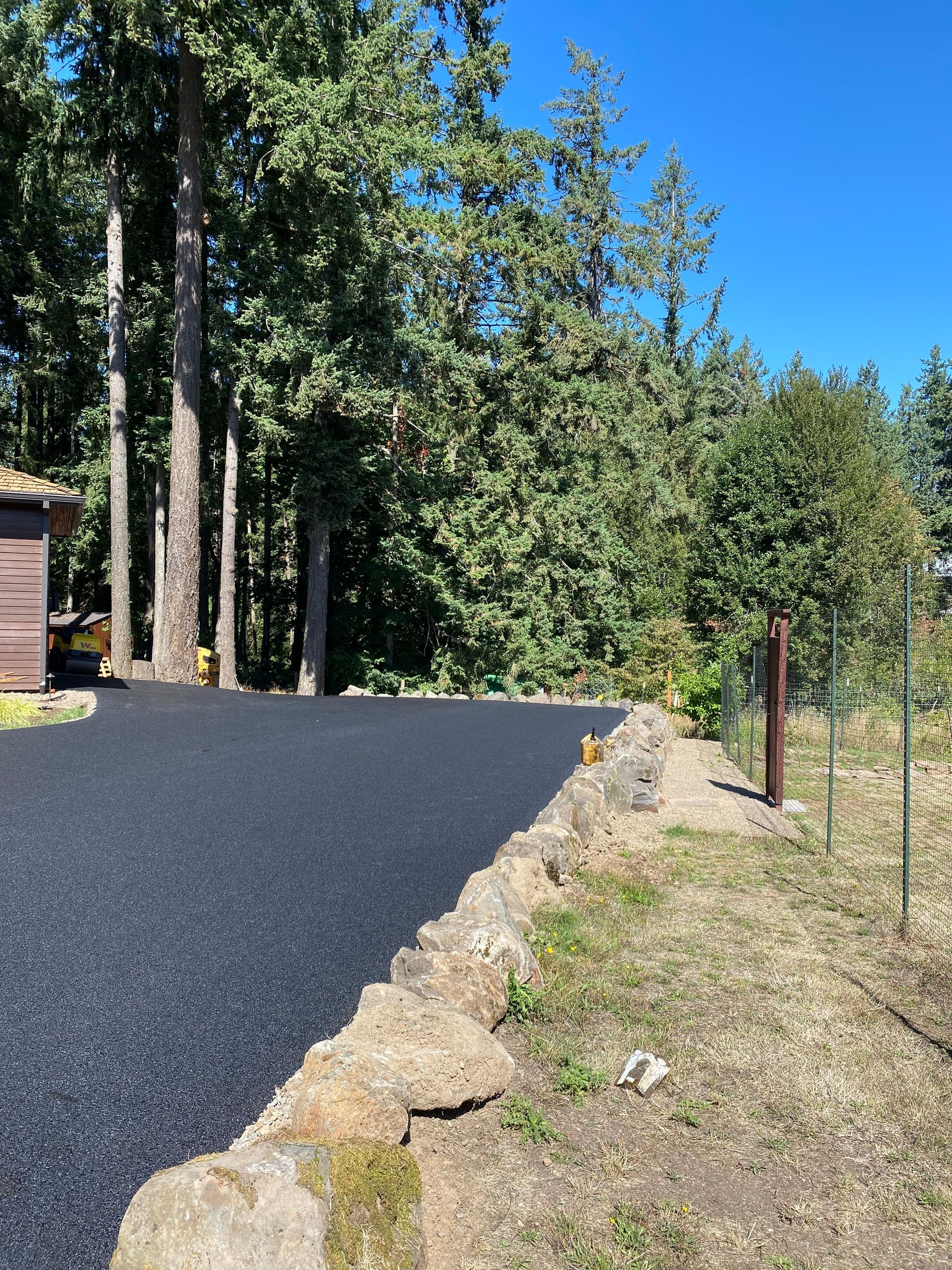 Black asphalt driveway alongside a stone retaining wall and a wire fence, with trees and a clear blue sky.