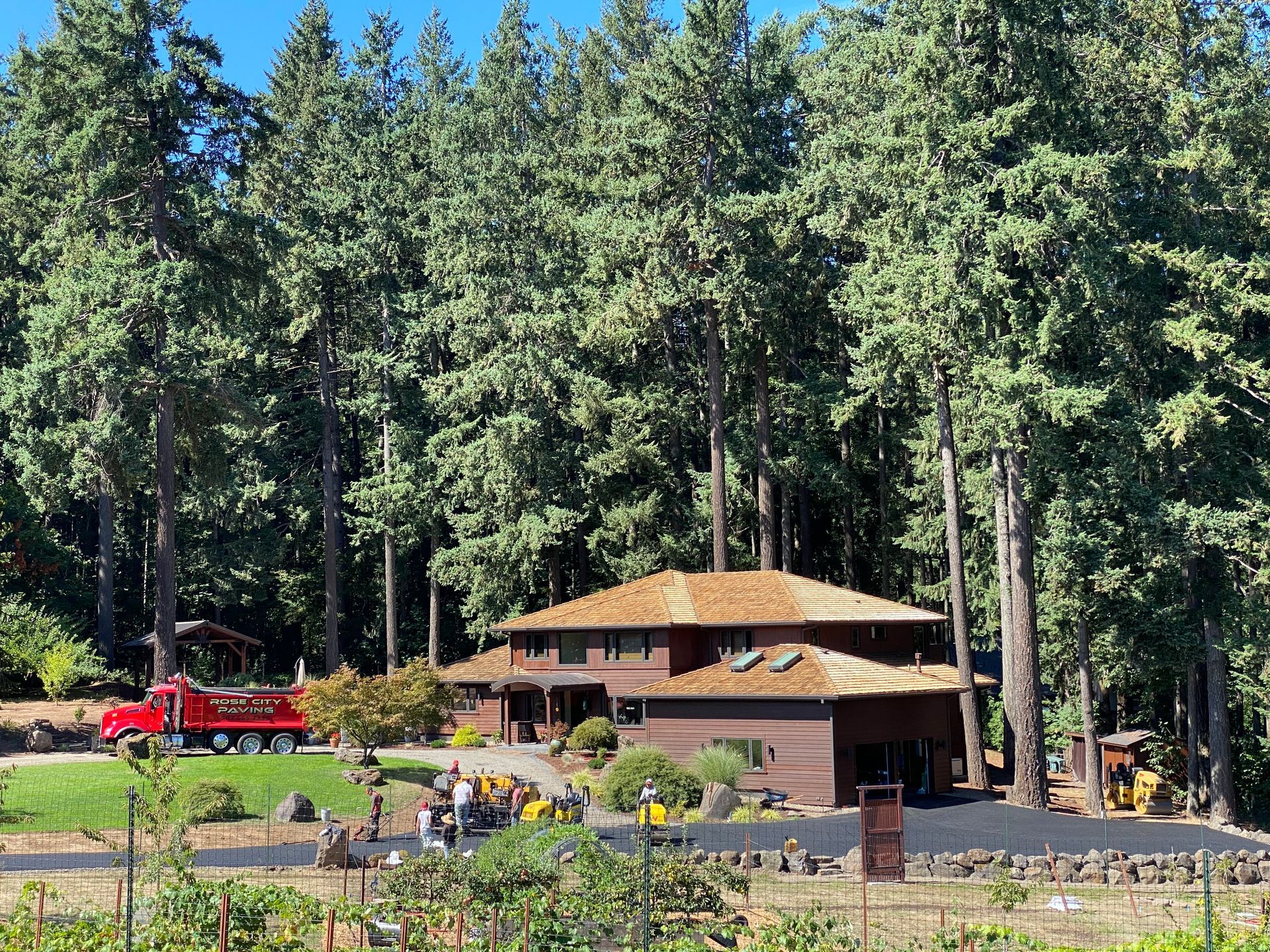 Brown house in front of tall green trees with a red truck, people, and equipment on the property.