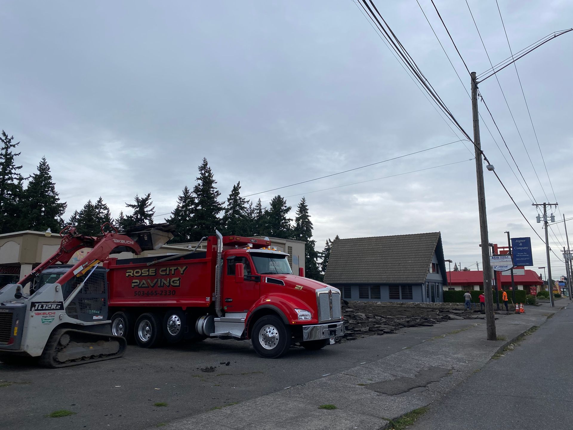 Red dump truck and excavator at a demolition site; cloudy sky overhead.
