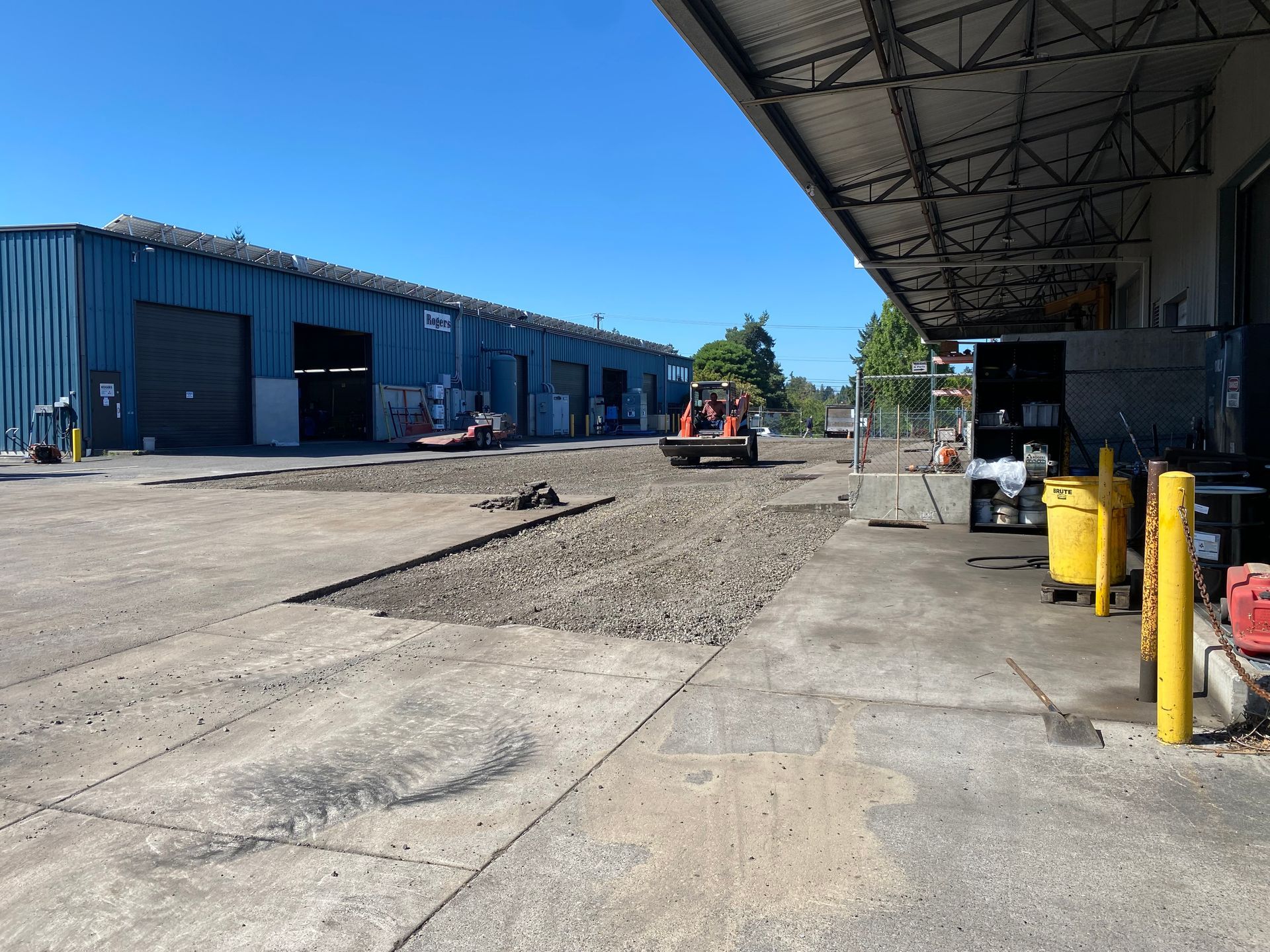 Construction site with a machine compacting gravel between two warehouse buildings on a sunny day.