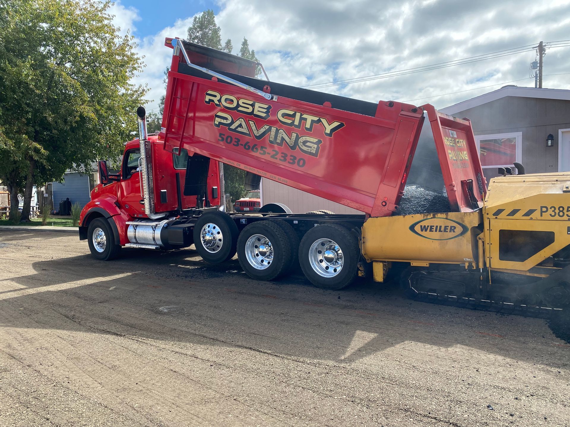 Red dump truck unloading asphalt into a paving machine; Rose City Paving.