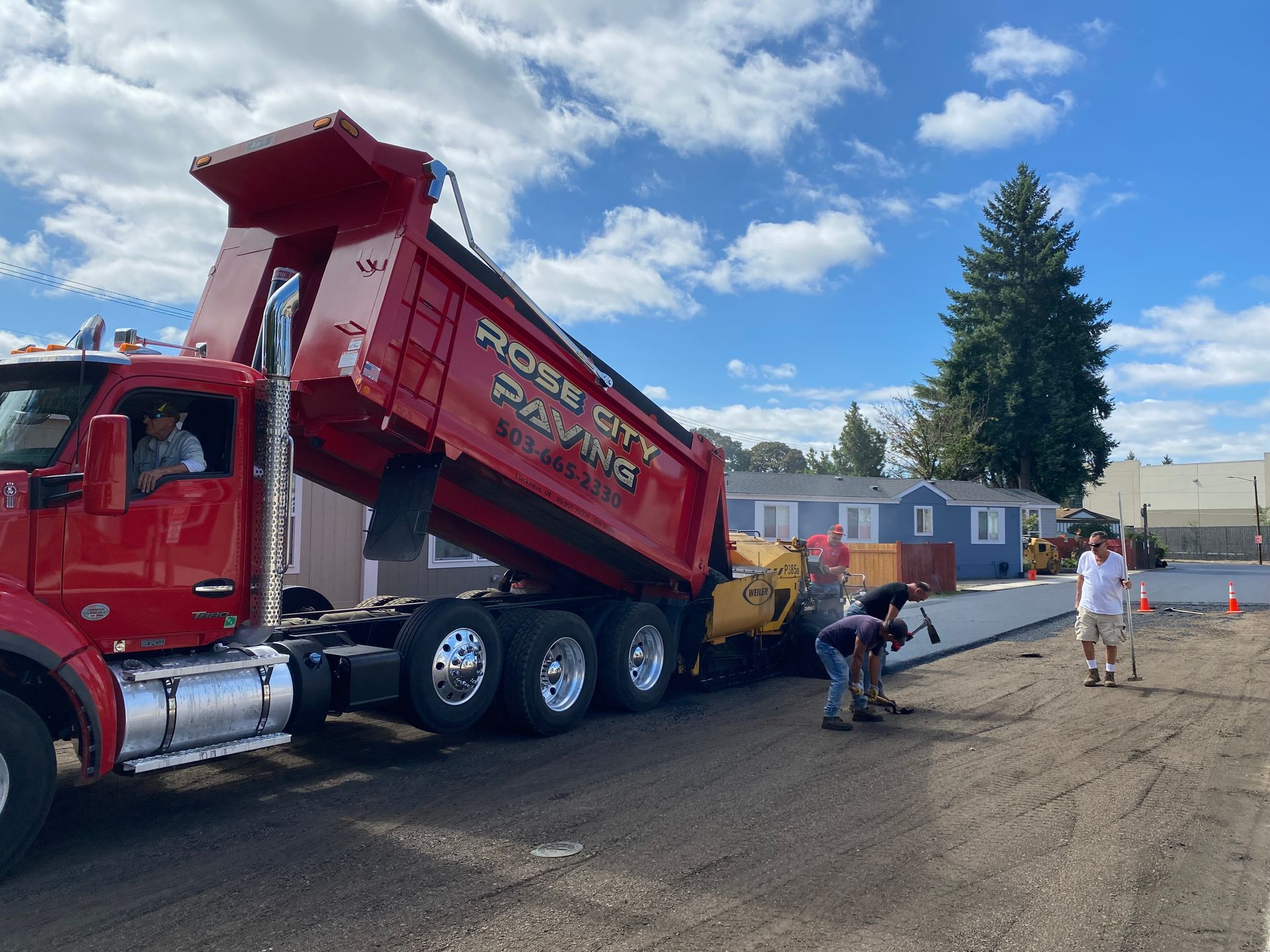 Red dump truck unloading asphalt for paving; workers and equipment present, blue sky.