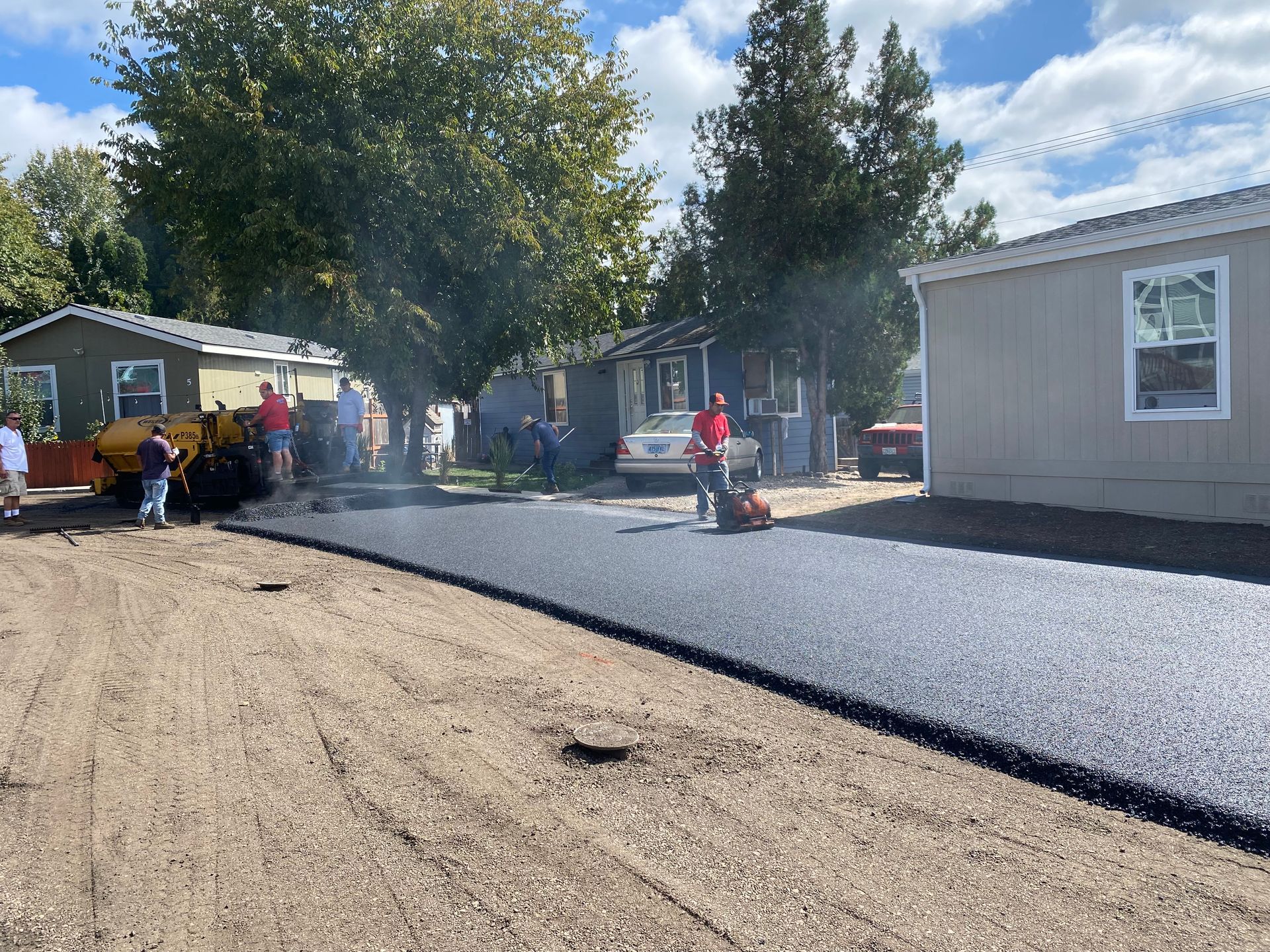 Asphalt road paving: workers and machinery paving a street lined with houses on a sunny day.