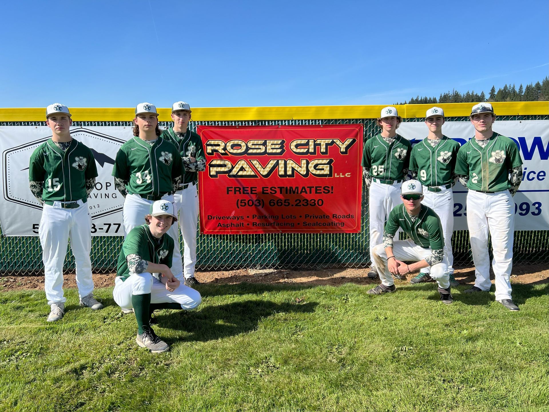 Baseball team posing on a field in front of a banner. Players wear green and white uniforms.