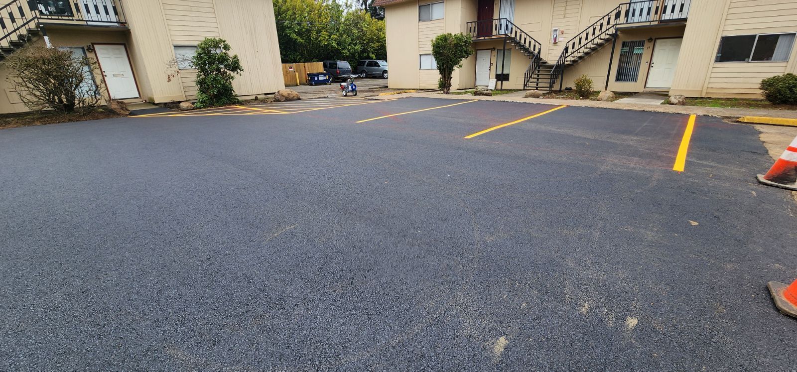 Newly paved asphalt parking lot with yellow painted lines and orange traffic cones. Apartment buildings in background.