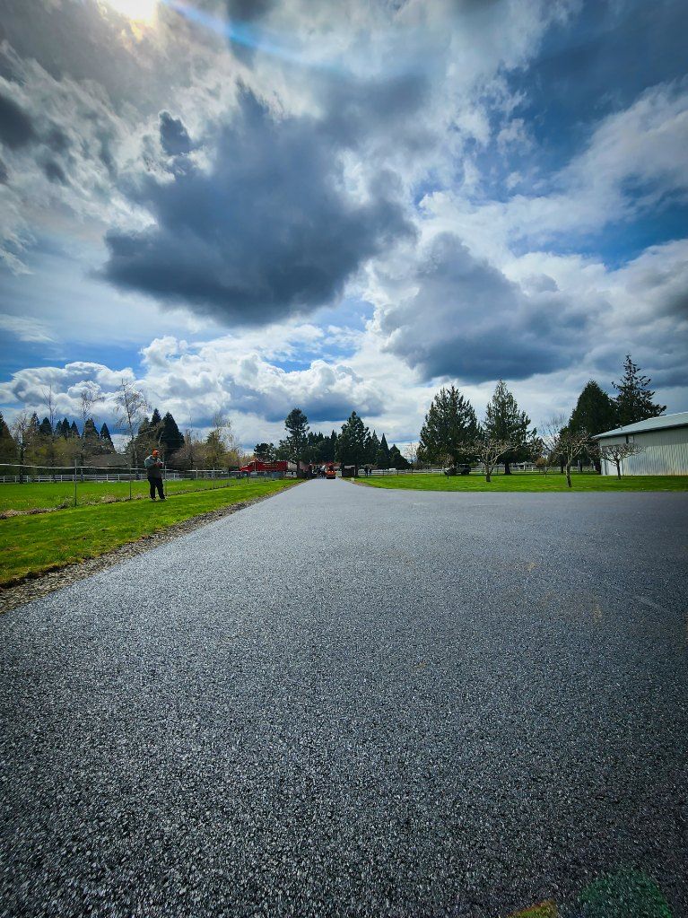 Paved road leads to trees under cloudy sky. Person in distance, green grass on sides.