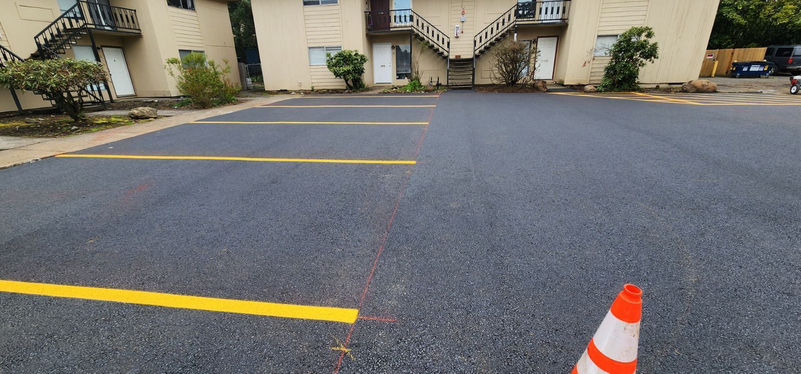 Newly paved parking lot with yellow painted lines and a traffic cone. Apartment building in the background.