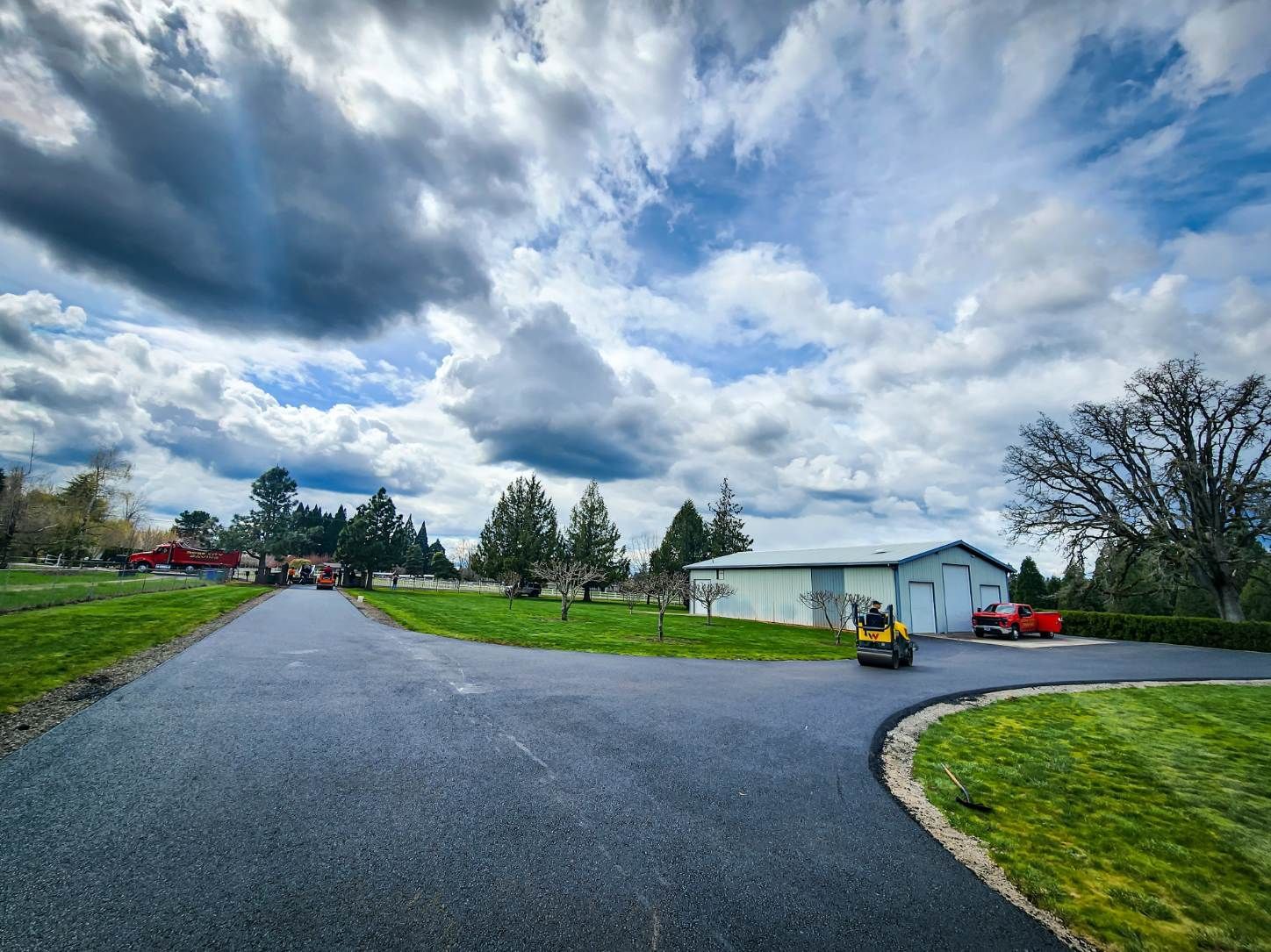 Newly paved asphalt driveway curves through a grassy landscape towards a building under a cloudy sky.