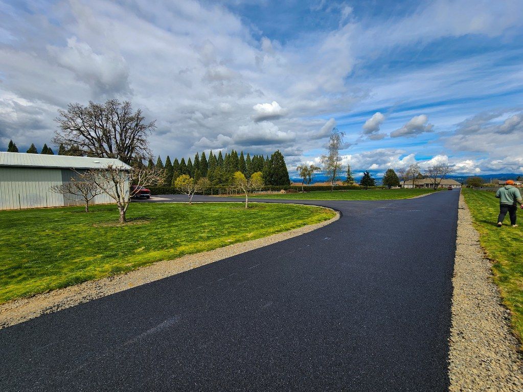 Asphalt driveway curves through grassy field, leading to trees and a barn under a cloudy sky.