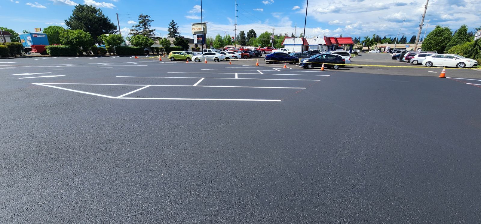 An empty parking lot with some cars parked in the distance. Blue sky and trees in background.