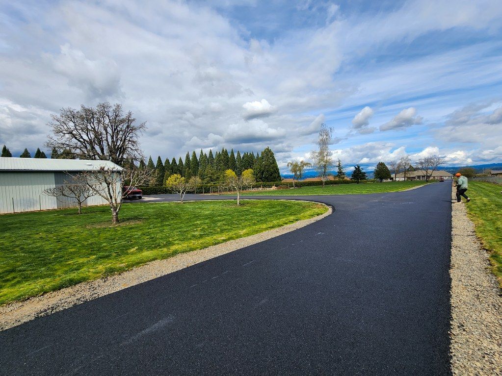 Paved driveway curves through green grass toward trees and buildings under a cloudy blue sky.