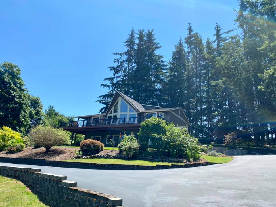 Brown house with a large deck, tall windows, and a sloped roof, surrounded by trees on a sunny day.