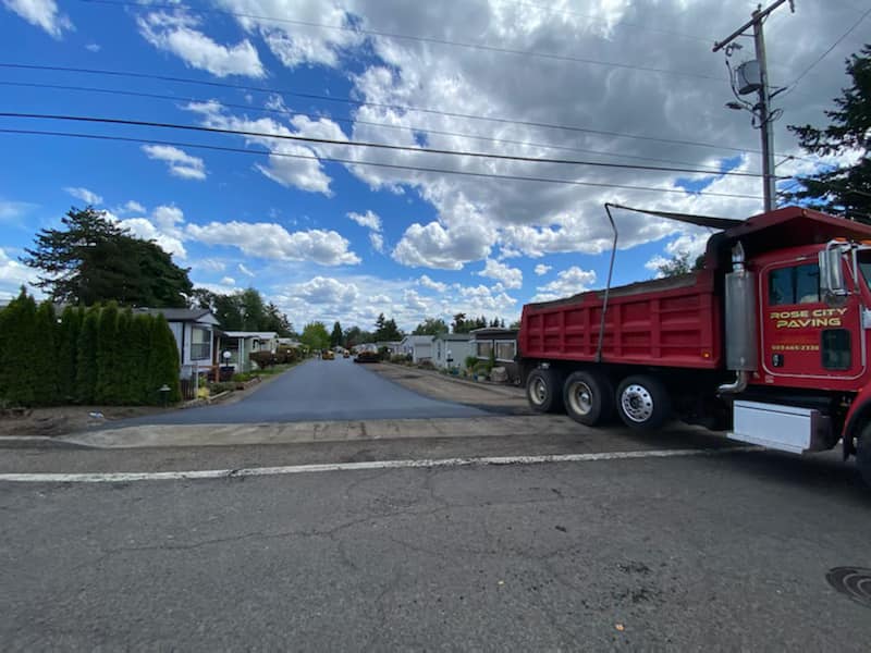 A red dump truck on a freshly paved road in a mobile home park under a cloudy sky.