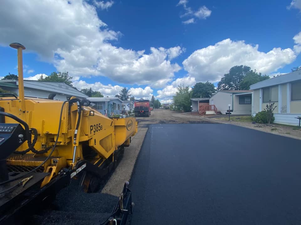 Asphalt paving in progress on a residential street; yellow paver, blue sky, clouds, and houses.