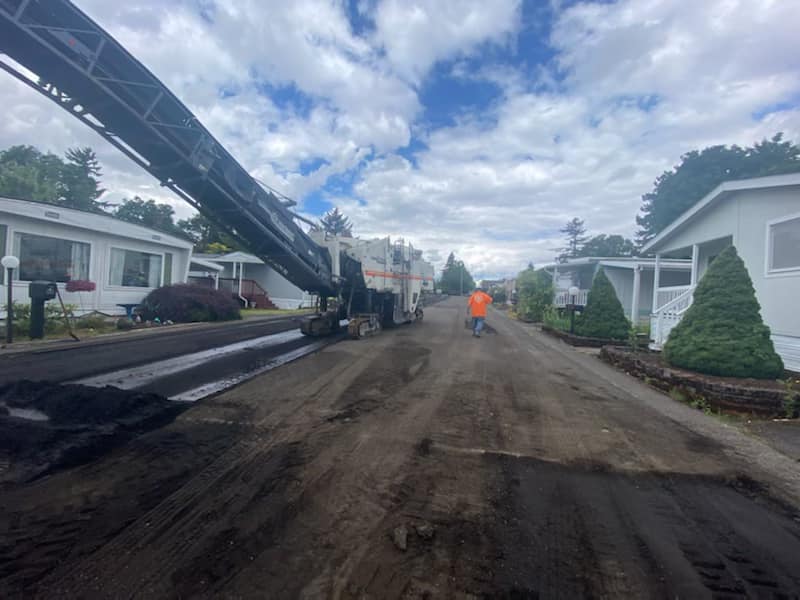 Road milling machine removing asphalt from a street in a mobile home park; worker in orange vest.