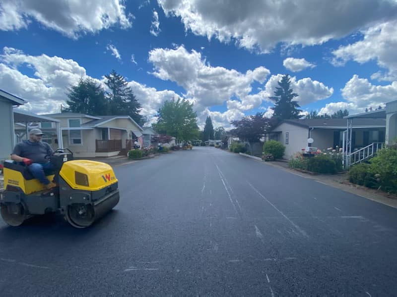A person operates a yellow roller compactor on a newly paved road in a residential area.