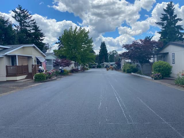 Street lined with mobile homes under a cloudy sky. Freshly paved road with some trees and flowers.