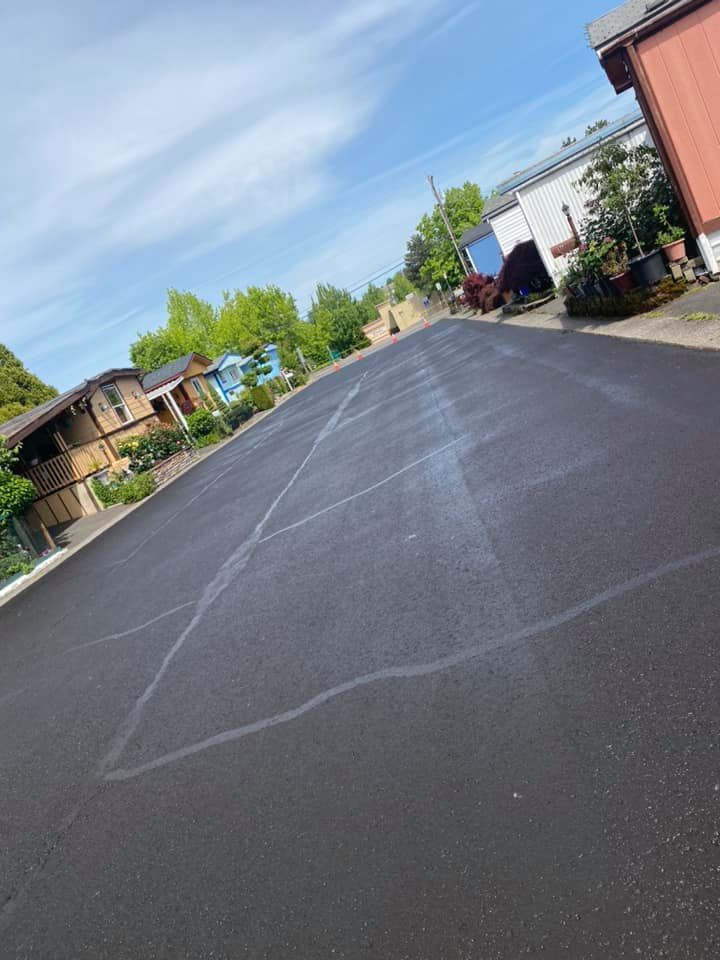 Newly paved asphalt road in a residential area, with houses and trees visible under a blue sky.