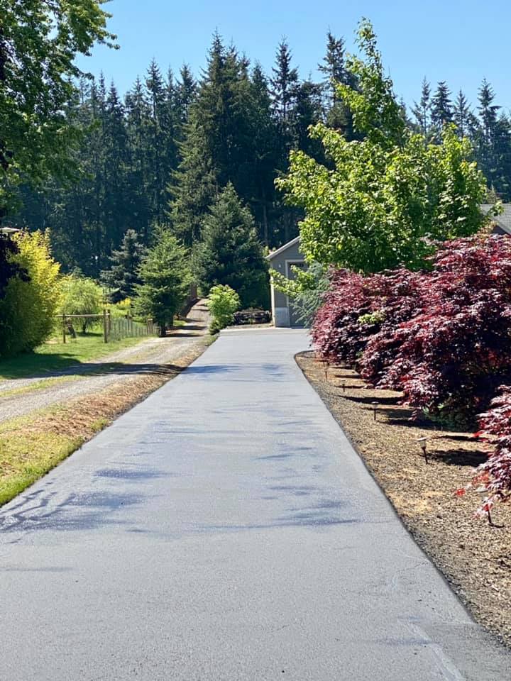Paved driveway lined with colorful bushes and trees, leading to a light gray building on a sunny day.
