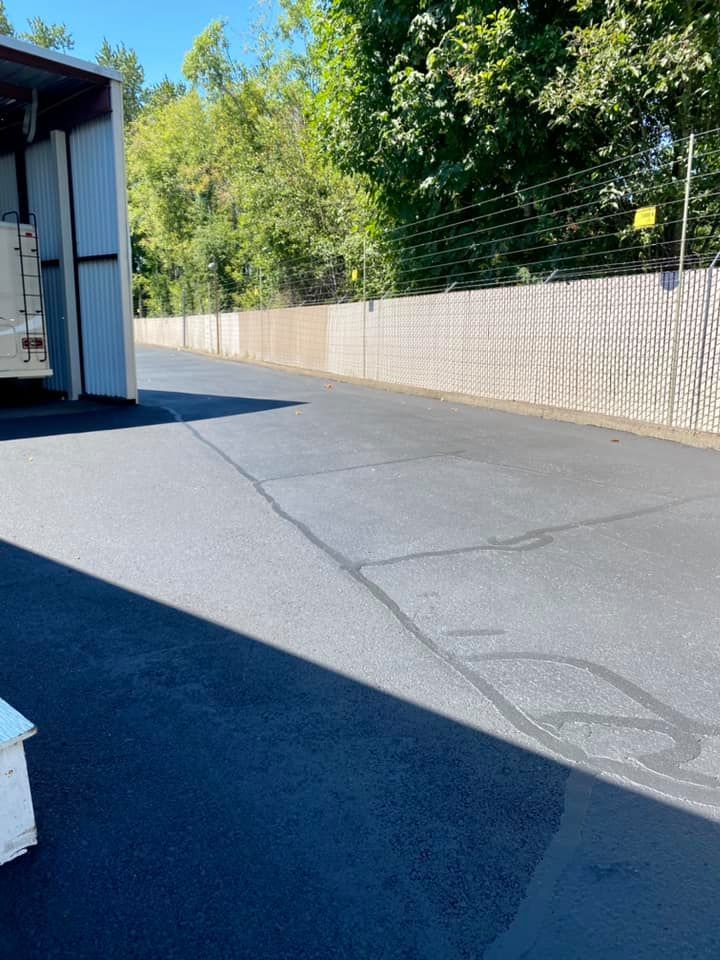 Asphalt driveway next to a light-colored fence. Trees and a building are in the background on a sunny day.