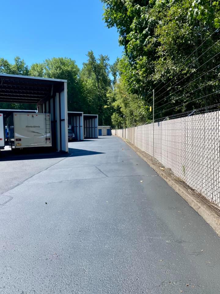 Driveway lined with covered storage units and chain link fence. Trees in background under a blue sky.