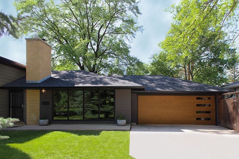 Modern single-story house with brick chimney, dark roof, large windows, and a wooden garage door beside a green lawn